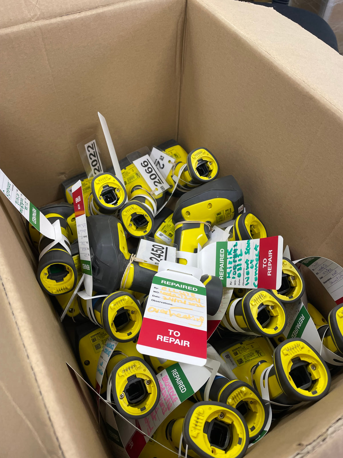A box of industrial barcode scanners with red TO REPAIR and green REPAIRED tags attached